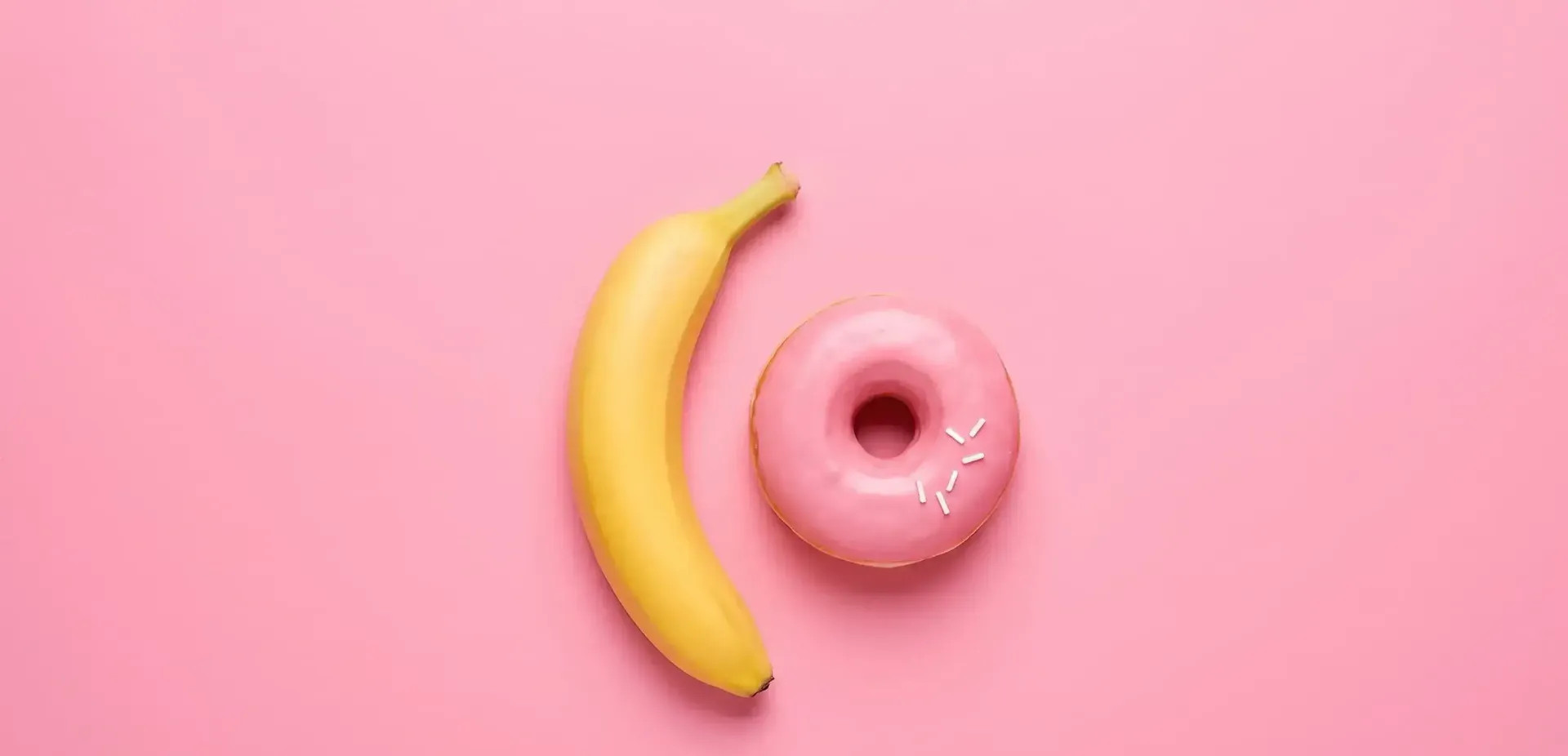 Hravá metafora pro orální sex – žlutý banán a růžový donut na růžovém pozadí.